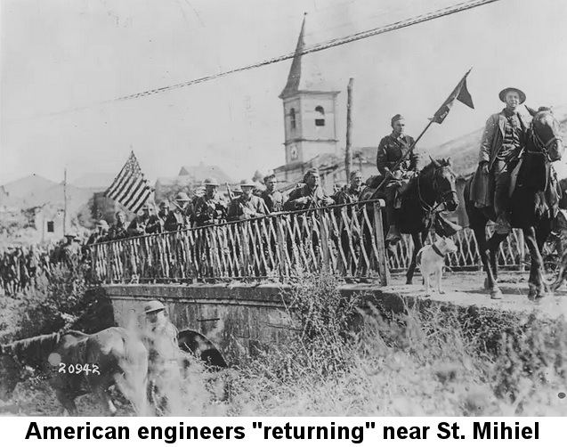 Black and white photo soldiers, one bearing an American flag, led by two officers on horseback, crossing a short bridge with a white church steeple in the background.